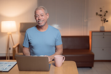 Happy retired man using laptop sitting at desk in his living room at night working from home with a...