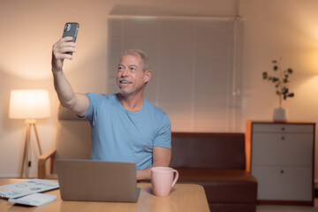 Smiling mature man taking a selfie with his smartphone while working from home at night, sitting at a table with his laptop and documents, enjoying a cup of coffee