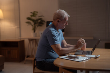 Worried senior man is sitting at a table in his living room at night, managing domestic finances and using a laptop, expressing concern and stress