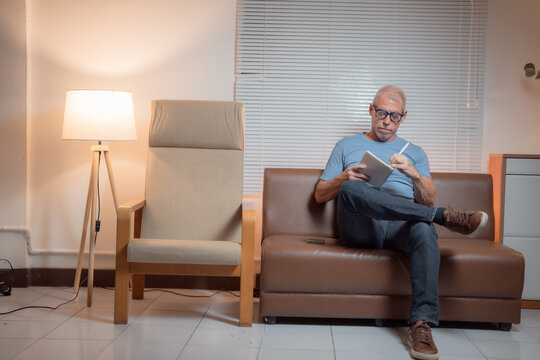 Focused mature man writing notes on notebook sitting on sofa in living room at home, with legs crossed and a serious expression, enjoying the quiet evening - Powered by Adobe