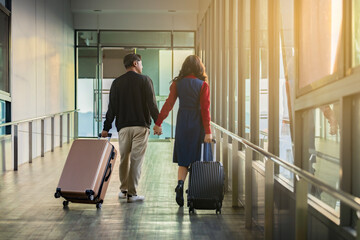 Back view of Happy Young asian couple with luggage walking hand in hand in an airport corridor signifies the joy of traveling together while going to boarding gate, Sunlight streams in. Copy Space