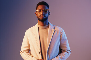 Stylish young African man wearing glasses and a light coat, standing against a colorful purple background, exuding confidence and modern fashion