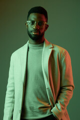Stylish male model with glasses in a green and red lit background, wearing a light coat and sweater, exuding confidence and modern elegance