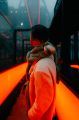 A woman in a warm coat and scarf stands on a brightly illuminated red escalator inside a futuristic glass structure. The atmosphere is vibrant, modern, and dynamic.