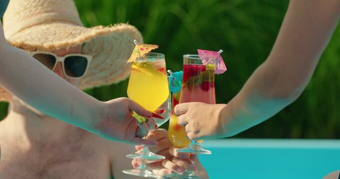 People toasting with colorful tropical cocktails by the swimming pool, garnished with fruit, mint, and decorative umbrellas, enjoying a refreshing summer moment.