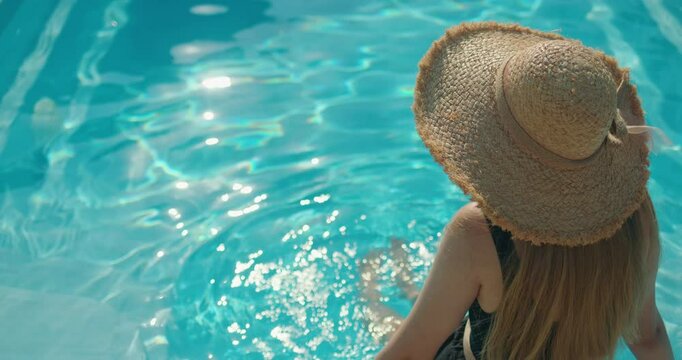Woman wearing a straw hat relaxing by a swimming pool, dipping her legs in the water on a sunny summer day.