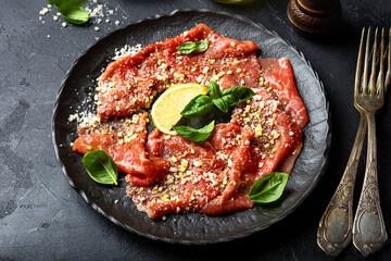 Slices of fresh beef carpaccio with basil and parmesan cheese in a black plate on the black background close up
