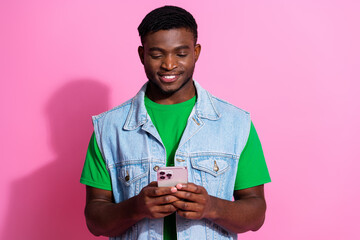 Young male in denim vest and green shirt using smartphone against vibrant pink background