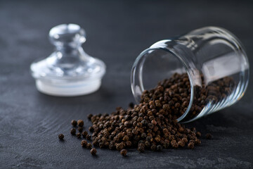 black peppercorns spill out of a glass storage jar on a black kitchen table, selective focus.