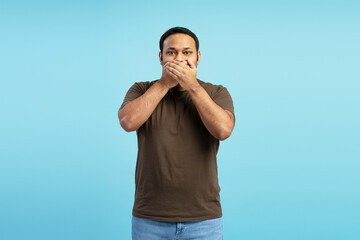 Middle aged Indian man covering his mouth on blue background