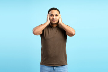 Young man covering ears ignoring noise on blue background