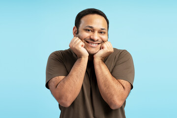 Enthusiastic man expressing excitement with fists under chin and big smile on blue background