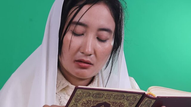A woman wearing a hijab is reading from a book quran with Arabic scrip, she has dark hair and is wearing white clothing, isolated on a green background or green screen