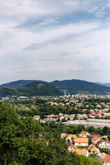 Obraz premium Scenic vista showcasing a cityscape nestled amidst lush greenery, with rolling hills stretching into the distance under a cloudy sky, offering a tranquil and picturesque view