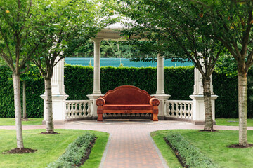 Elegant red sofa sits beneath a white pergola in a meticulously maintained garden. Offering a peaceful retreat amidst lush greenery and manicured hedges. Creating a serene and inviting atmosphere