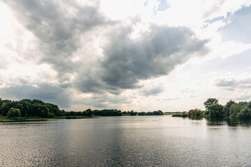Dark storm clouds hovering above expansive river, verdant banks framing brooding natural scenery...
