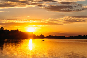 Small boat crossing calm lake water during a breathtaking golden hour sunset, creating a beautiful reflection on the water's surface with city silhouette on background