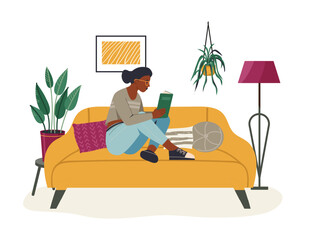 Fototapeta premium Young black woman is reading a book sitting on a comfortable yellow sofa in her living room, enjoying her free time surrounded by houseplants and a modern floor lamp
