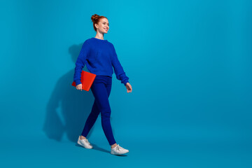 Young stylish woman in casual blue outfit holding a book against bright blue background, expressing cheerful and confident attitude
