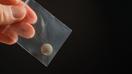 Hand of the drug dealer show off and lure with prescription opioid pill in small plastic bag for sale in clubs, parties and street to addicts. Illegal drugs trafficking, cause dependence and overdose