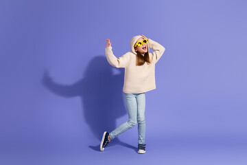 Cheerful young girl in casual wear and sunglasses posing on a vibrant purple studio background