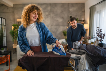 A woman iron shirts on an ironing board, while a man folds up them