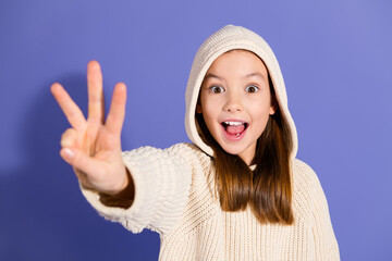 Charming young girl in a casual hoodie smiling joyfully against a purple background, holding up peace sign