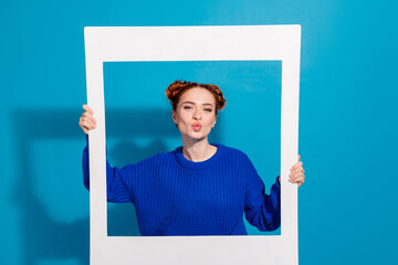 Young red-haired woman in blue sweater making a playful gesture holding a frame against a bright...
