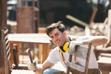 Male carpenter using hammer repairing wooden arm chair at woodwork workshop. Male joiner working...