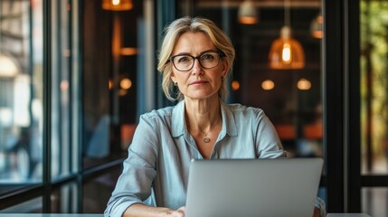 Busy middle aged business woman executive wearing shirt and glasses sitting at desk using laptop on glass big office . Mature serious professional female manager working looking at computer technology