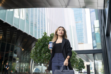A confident young professional woman walking outdoors in a modern urban setting, holding a coffee...