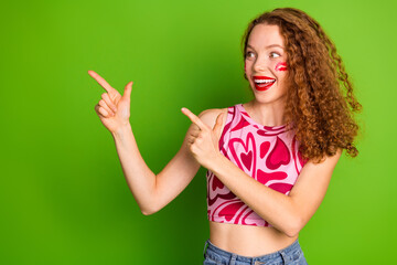 Obraz premium Joyful young woman with curly red hair pointing upwards against vibrant green background