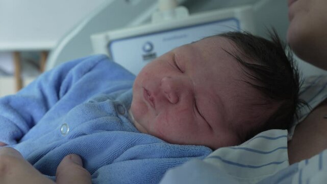 Close-up of a newborn baby in a blue onesie, peacefully sleeping on a parent's chest, highlighting the deep bond and the serene moments of early infancy in a hospital setting