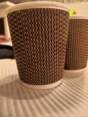 A close-up of a brown textured disposable coffee cup placed on a wooden table. The cup has an intricate woven-like pattern, giving it a unique and stylish appearance.


