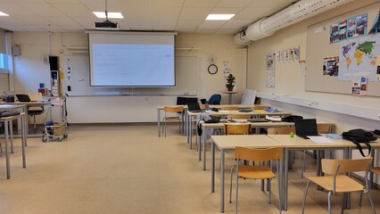 The classroom features multiple desks arranged for students with a projector screen at the front....