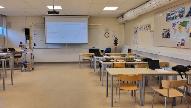 The classroom features multiple desks arranged for students with a projector screen at the front. The space is well-lit and prepared for an upcoming lesson with educational materials around.