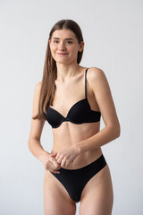 Fototapeta premium young woman with long hair stands in front of a white background, wearing black lingerie and black heels. She looks directly at the camera with a slight smile. Minimalistic studio lighting, natural 