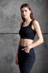 Fototapeta premium A professional modeling snapshot of a young female model in a black crop top and high-waisted jeans, posing against a textured gray wall. Capturing her natural elegance, toned physique, and confident 