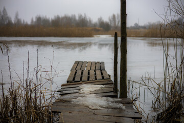 Obraz premium Old wooden pier covered with patches of ice extends into a partially frozen lake surrounded by dry reeds and bare trees in a bleak winter landscape. Old bridge on the pond.