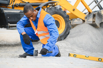 African american master in gloves lays paving stones in layers. Laying gray concrete paving slabs in house courtyard on sand foundation base