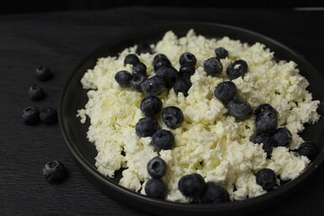 The cottage cheese in the black plate. On a black background. Close up of cottage cheese with blueberries. Honey watering curd on black background. 