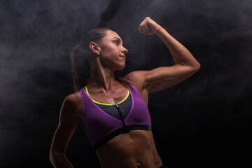 Obraz premium Confident woman flexing bicep in sporty attire against a dark smoky background