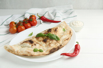 Tasty calzone, basil, chili pepper, sauce and tomatoes on white wooden table, closeup