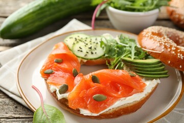 Delicious bagel with salmon, cream cheese, cucumber, avocado and pumpkin seeds on wooden table, closeup