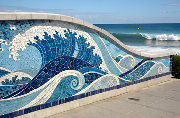 A vibrant mosaic mural of ocean waves in blues and whites, set against a beach backdrop with gentle waves lapping at the shore.