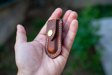 handcrafted brown leather knife sheath with a brass snap button in a man hand featuring detailed stitching and a metal loop, against a blurred green outdoor background