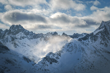 Wind sweeps snow over Sattelgrat and Sulzspitze, illuminated by sunlight. Verwall mountains, including Madaunspitze, Rendlspitze, and Riffelspitze, form the background.
