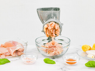 Manual retro meat grinder with minced meat in a glass bowl, boneless chicken thighs, pink salt, peeled onions, garlic in a saucer and basil