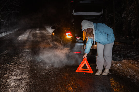 On a snowy night road, a woman places a warning triangle behind her broken-down car in a winter forest. Steam rises from the vehicle, highlighting the cold, remote environment.