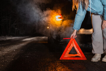 On a dark winter road, a woman places an emergency triangle behind her broken-down car. The scene...